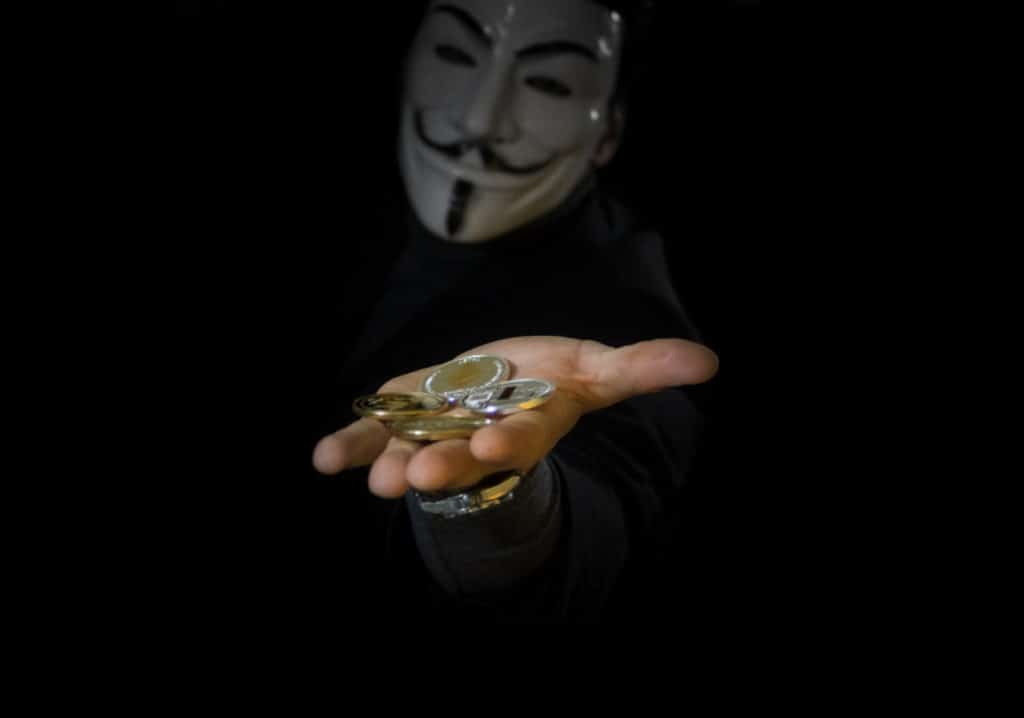Uma mão estendida segurando moedas vindas de uma pessoa usando uma máscara 'anônima'.